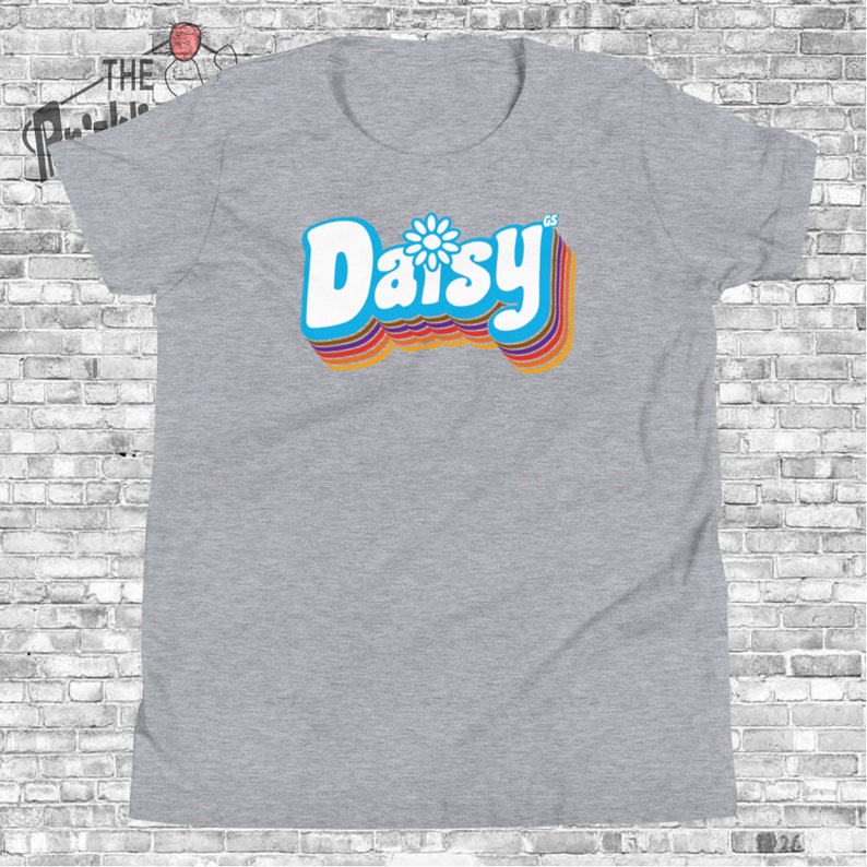 May include: A gray t-shirt with a retro-style graphic of the word "Daisy" in blue with a white daisy in the center. The word is surrounded by rainbow-colored stripes.