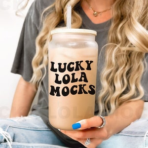 May include: A clear glass tumbler with a white lid and a black straw. The tumbler is filled with a light brown beverage and has the text "LUCKY LOLA MOCKS" printed on the side.