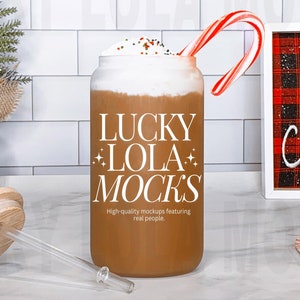 Pode incluir: Uma caneca de cer&acirc;mica branca com um l&iacute;quido castanho e uma cobertura de chantilly. A caneca tem o texto "LUCKY LOLA MOCKS" impresso nela. A caneca est&aacute; sentada em uma bancada branca com um bast&atilde;o de caramelo, uma tampa de madeira e um canudo transparente. Uma placa emoldurada com um fundo xadrez vermelho e preto diz "LET'S GET Cozy".