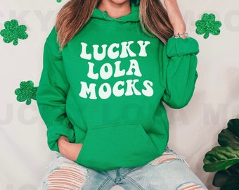 May include: A green hoodie with the text "LUCKY LOLA MOCKS" in white. The hoodie is worn by a person with long brown hair and is sitting down.