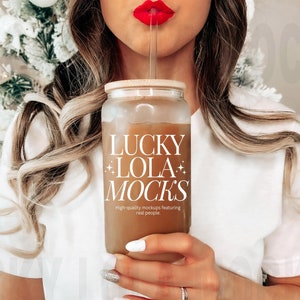 May include: A woman holds a clear glass tumbler with a brown liquid and a straw. The tumbler has the text "LUCKY LOLA MOCKS" and "High-quality mockups featuring real people." printed on it.