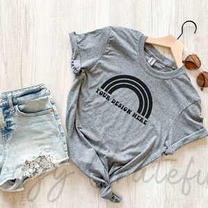 May include: A gray t-shirt with a black rainbow graphic and the text "Your Design Here". The t-shirt is tied in a knot at the bottom and is laying on a wooden surface. There are also a pair of denim shorts and sunglasses in the image.