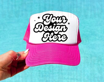 May include: A pink and white trucker hat with a black and white retro font design that says "Your Design Here" with two white stars on either side of the text.