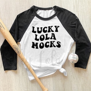 May include: A white and black baseball tee shirt with the text "Lucky Lola Mocks" in black. A wooden baseball bat and a white baseball are on a wooden surface.
