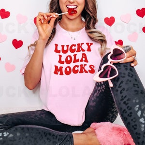 May include: A pink t-shirt with the text "LUCKY LOLA MOCKS" in red letters. The person wearing the shirt is holding a lollipop and wearing black leggings with a leopard print.