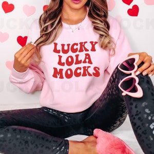 May include: A pink sweatshirt with the text "LUCKY LOLA MOCKS" in red letters. The model is wearing black leggings with a leopard print and pink fuzzy slippers.