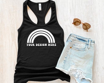 May include: Black racerback tank top with a white rainbow graphic and the text "YOUR DESIGN HERE". The tank top is laying on a light wood surface with a pair of light blue denim shorts and brown sunglasses.