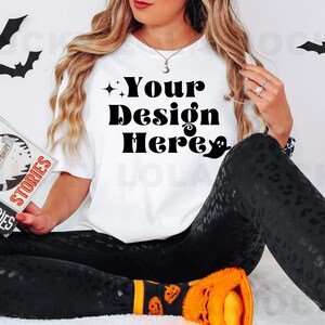 May include: A white t-shirt with the text "Your Design Here" in black lettering. The shirt is being worn by a person with long blonde hair and black pants with a leopard print. The person is holding a book with the title "Ghost Stories" in their hands.