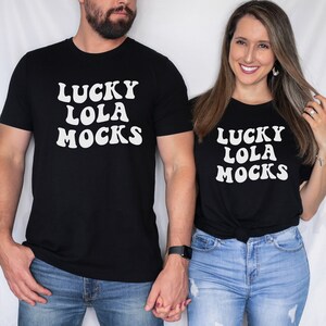 Puede incluir: Una pareja lleva camisetas negras a juego con texto blanco que dice "Lucky Lola Mocks". El hombre lleva pantalones vaqueros azules y la mujer lleva pantalones vaqueros azules con detalles desgastados.
