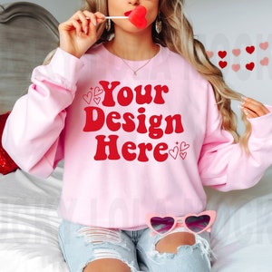 May include: A pink sweatshirt with the text "Your Design Here" in red letters. The sweatshirt has a crew neck and long sleeves. The model is wearing the sweatshirt with a pair of ripped denim shorts.