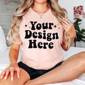 Peut inclure: Un t-shirt rose avec le texte "Your Design Here" en lettres noires de style rétro. Le t-shirt est porté par une personne assise sur un lit.