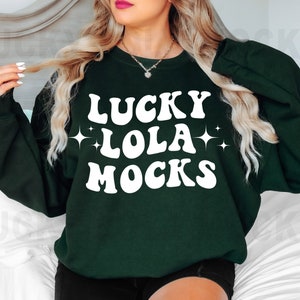 May include: A dark green sweatshirt with white text that reads "LUCKY LOLA MOCKS" in a retro font. The text is surrounded by two white stars.