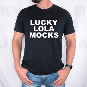 May include: A black t-shirt with white text that reads "LUCKY LOLA MOCKS". The person is wearing blue jeans.