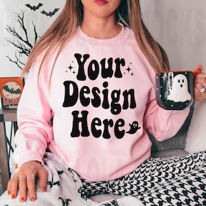 May include: A pink sweatshirt with the text "Your Design Here" in black retro font. The sweatshirt is worn by a person sitting on a couch with a black and white patterned blanket.