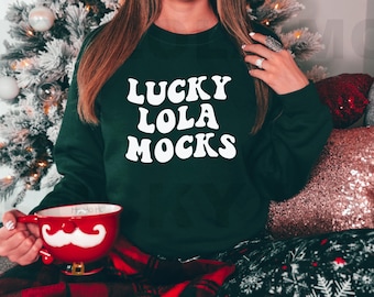May include: A woman wearing a green sweatshirt with the text "LUCKY LOLA MOCKS" in white. She is sitting on a couch with a red and black plaid blanket and a red mug with a white mustache.
