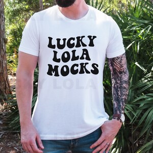 以下が含まれることがあります： レトロなフォントで「LUCKY LOLA MOCKS」と書かれた黒いテキストがプリントされた白いTシャツ