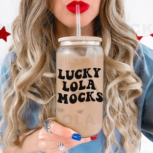 May include: A woman with long blonde hair is drinking from a clear glass with a brown lid. The glass has the text "LUCKY LOLA MOCKS" printed on it in black.