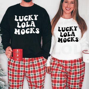 Puede incluir: Una pareja lleva sudaderas a juego en blanco y negro con el texto "Lucky Lola Mocks" en una fuente retro. Ambos llevan pantalones de pijama a cuadros rojos y verdes.