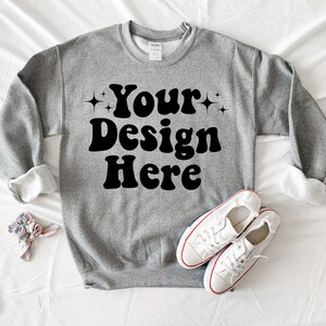 May include: A grey sweatshirt with the text "Your Design Here" in black retro-style lettering. The sweatshirt is laid flat on a white surface with a pair of white trainers.