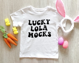 May include: White baby t-shirt with black text that reads "LUCKY LOLA MOCKS". The shirt is laying on a wooden surface with Easter decorations, including yellow chicks, carrots, and pink eggs.