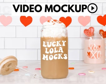 May include: A clear glass tumbler with a brown beverage and white whipped cream topping. The tumbler has a white sticker with the text "LUCKY LOLA MOCKS". The tumbler is on a white countertop with a wooden lid and a glass straw.