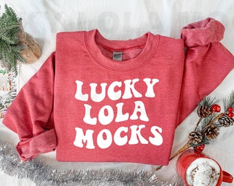 May include: Red sweatshirt with white text that reads "Lucky Lola Mocks". The sweatshirt is folded and laid on a white surface with silver tinsel, a small potted evergreen plant, a brown burlap wrapped gift, and a red mug with a white beverage.