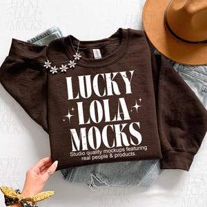 Könnte beinhalten: Ein dunkelbraunes Sweatshirt mit dem weißen Text "LUCKY LOLA MOCKS", einer Blumenkette, einem braunen Hut und einer Jeans. Das Sweatshirt hat den Text "Studio quality mockups featuring real people & products."