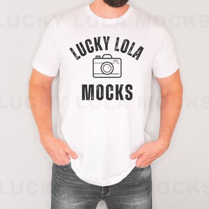 May include: White t-shirt with the text "LUCKY LOLA MOCKS" in black, with a camera graphic in the center. The shirt is being worn by a person with dark hair and a beard, wearing blue jeans.