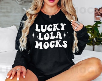Op de afbeelding: Een zwart sweatshirt met de tekst "LUCKY LOLA MOCKS" in witte retro-lettertype met glinsterende sterren aan weerszijden van het woord "LOLA".