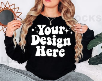 Op de afbeelding: Een zwart sweatshirt met de tekst "Your Design Here" in witte retro-stijl letters. Het sweatshirt wordt gemodelleerd door een persoon met lang blond haar.