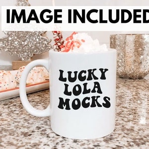 May include: White ceramic mug with black text that reads "LUCKY LOLA MOCKS". The mug is filled with a white beverage topped with whipped cream. The mug is on a countertop with a festive background.