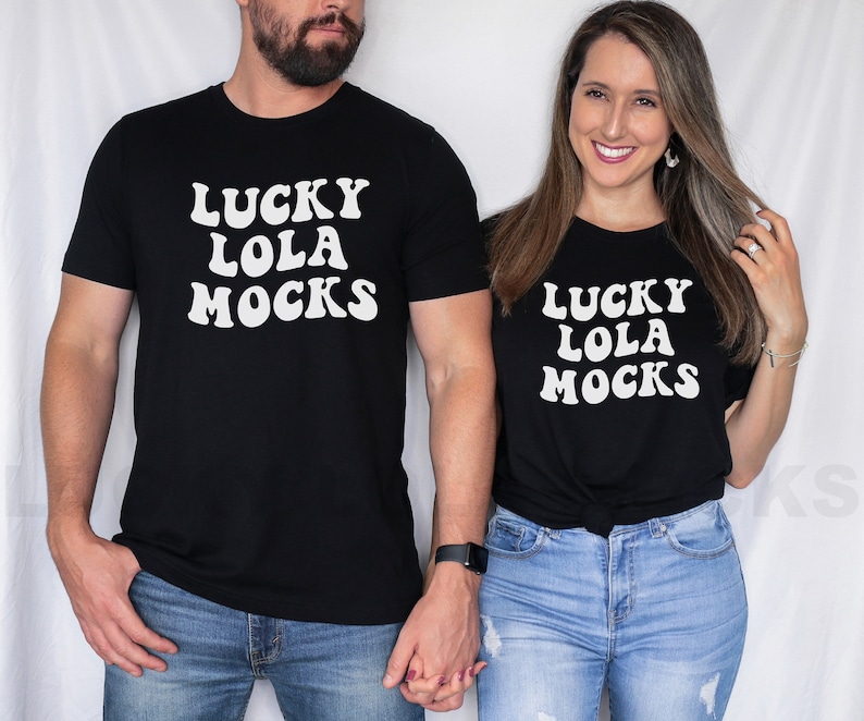 Puede incluir: Una pareja lleva camisetas negras a juego con texto blanco que dice "Lucky Lola Mocks". El hombre lleva pantalones vaqueros azules y la mujer lleva pantalones vaqueros azules con detalles desgastados.