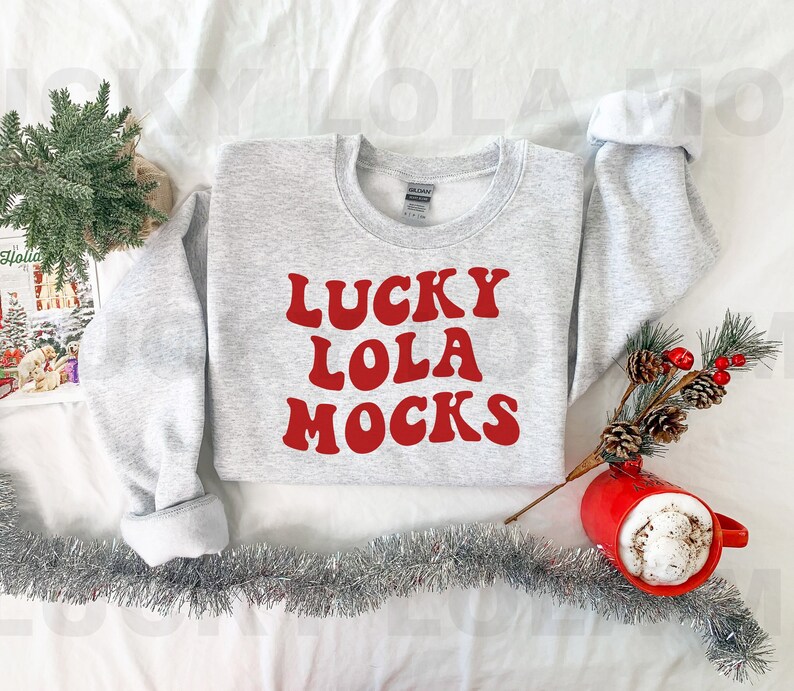 Op de afbeelding: Een grijs sweatshirt met de tekst "LUCKY LOLA MOCKS" in rode letters. Het sweatshirt ligt op een wit oppervlak met zilveren tinsel en een rode mok met een witte drank.
