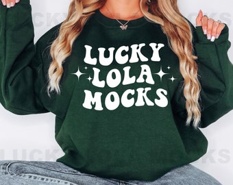 May include: A dark green sweatshirt with white text that reads "Lucky Lola Mocks" in a retro font. The text is surrounded by small white stars.