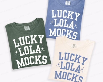 May include: Three folded t-shirts in different colors: green, blue, and cream. Each shirt has the text "LUCKY LOLA MOCKS" in a bold, collegiate-style font. The text is white on the green shirt, and blue on the cream shirt. The blue shirt has white text.