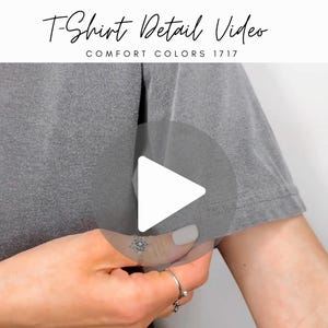 May include: A close-up of a gray t-shirt with the text "T-Shirt Detail Video" and "Comfort Colors 1717". A white play button is overlaid on the shirt. The person is wearing rings on their fingers.