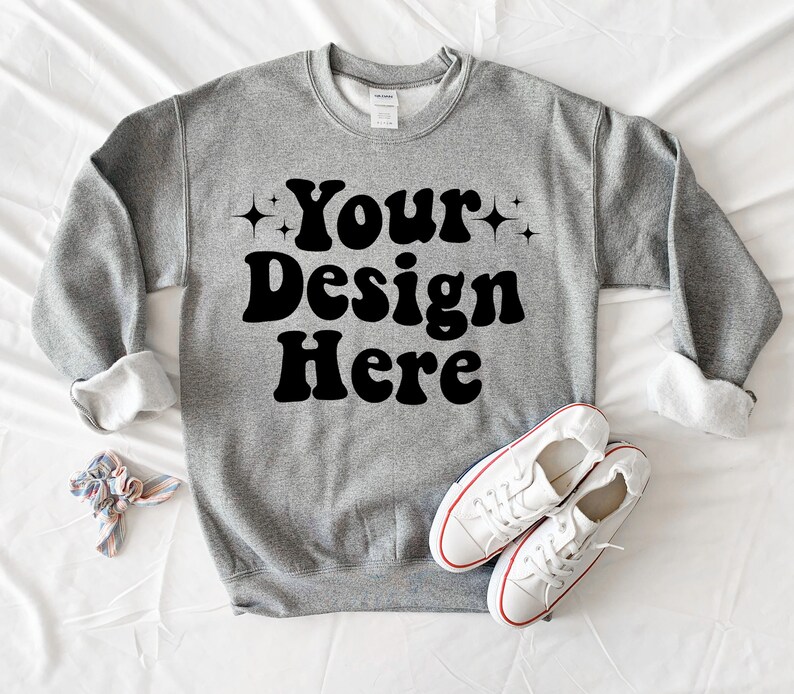 May include: A grey sweatshirt with the text "Your Design Here" in black retro-style lettering. The sweatshirt is laid flat on a white surface with a pair of white trainers.