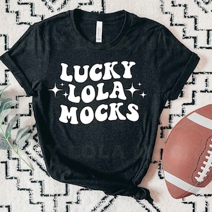 May include: Black t-shirt with white text that reads "Lucky Lola Mocks" in a retro font. The shirt is on a white and black patterned surface with a brown football.