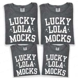 May include: Four gray t-shirts with the text "LUCKY LOLA MOCKS" in white block letters. The text includes star accents. The shirts are folded and stacked, showcasing the design on the front.