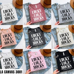 May include: A collection of Bella Canvas 3001 t-shirts in various colors, including white, maroon, gray, peach, black, and pink. Each shirt features the text "LUCKY LOLA MOCKS" in a bold, stylized font. The shirts are displayed with jeans and a brown hat.
