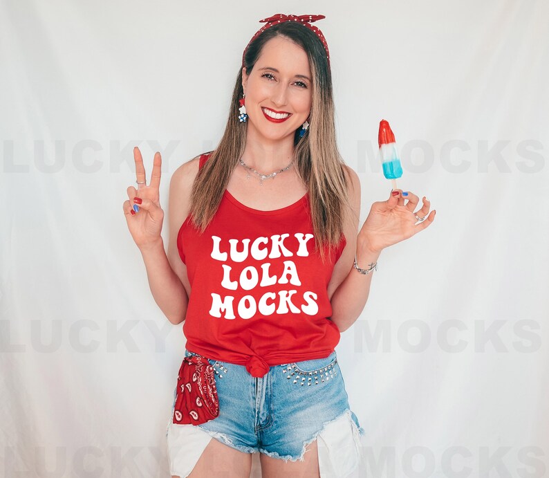 K&ouml;nnte beinhalten: Eine Frau tr&auml;gt ein rotes Tanktop mit dem Text "Lucky Lola Mocks" in Wei&szlig;. Sie h&auml;lt ein rot-wei&szlig;-blaues Eis am Stiel und tr&auml;gt ein rot-wei&szlig;-blaues Stirnband.