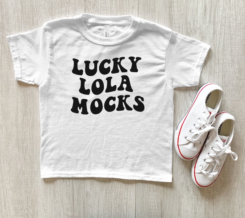 May include: White t-shirt with black text that reads "LUCKY LOLA MOCKS" in a retro font style. The shirt is laying on a wood surface with a pair of white sneakers next to it.