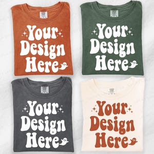Pode incluir: Quatro camisetas dobradas em ferrugem, verde escuro, cinza carvão e creme. Cada camiseta apresenta o texto "Your Design Here" em uma fonte retrô, com estrelas e um fantasma. Roupas para designs personalizados.
