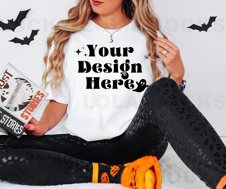 May include: A white t-shirt with the text "Your Design Here" in black lettering. The shirt is being worn by a person with long blonde hair and black pants with a leopard print. The person is holding a book with the title "Ghost Stories" in their hands.