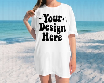 May include: White oversized t-shirt with the text "Your Design Here" in black retro font. The shirt is worn by a person standing on a sandy beach.