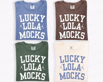 May include: Four folded t-shirts in different colors: blue, brown, green, and cream. Each shirt has the text "LUCKY LOLA MOCKS" in white or blue, with star accents. The shirts are made of a soft material.