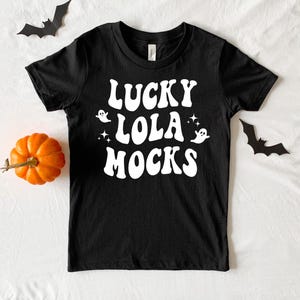 May include: A black short-sleeve t-shirt featuring white text that reads LUCKY LOLA MOCKS in a wavy, retro-inspired font. The design includes two small white ghost illustrations and two four-pointed star icons positioned around the lettering.