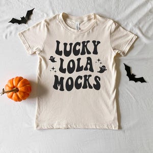 Könnte beinhalten: Ein cremefarbenes Kurzarm-T-Shirt mit schwarzem Text, der in einer wellenförmigen Schrift LUCKY LOLA MOCKS lautet. Das Design enthält kleine schwarze Geister- und Sterngrafiken. Das T-Shirt ist von einem kleinen orangefarbenen Kürbis und zwei schwarzen Papierfledermäusen umgeben.