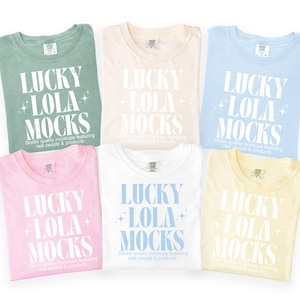 May include: Six pastel-colored t-shirts are displayed, each folded and showcasing the text "LUCKY LOLA MOCKS" in a bold, white font. The shirts are in shades of green, cream, blue, pink, white, and yellow. The text "Studio quality mockups featuring real people & products" is also visible.