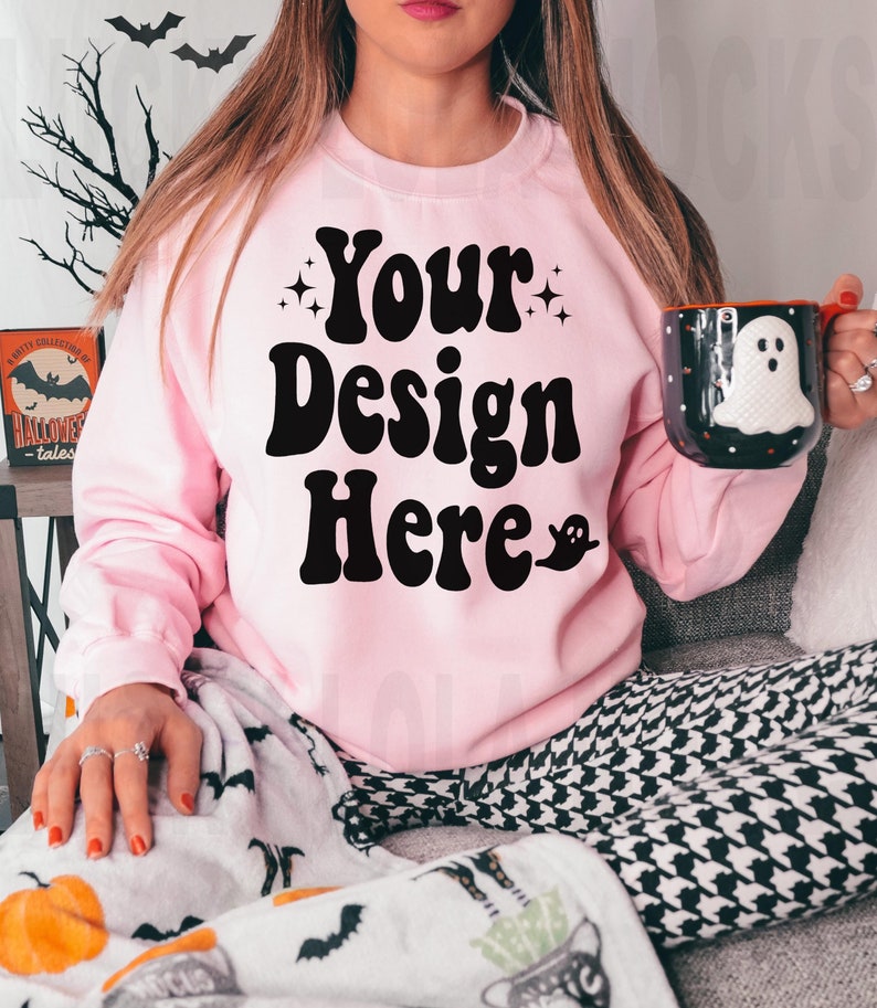 May include: A pink sweatshirt with the text "Your Design Here" in black retro font. The sweatshirt is worn by a person sitting on a couch with a black and white patterned blanket.